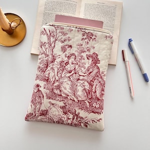 May include: A cream-coloured, quilted zippered pouch with a red toile print showing a pastoral scene with figures. The rectangular pouch, which appears to be holding a book, is accompanied by two pens and a book.