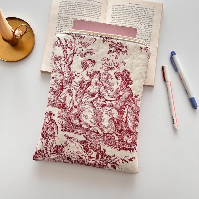 May include: A cream-coloured, quilted zippered pouch with a red toile print showing a pastoral scene with figures. The rectangular pouch, which appears to be holding a book, is accompanied by two pens and a book.