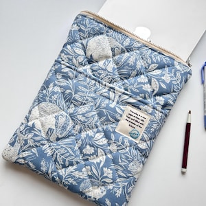 May include: Blue quilted laptop sleeve with a white floral and hedgehog pattern. The sleeve has a zipper closure and a small tag with text. A pen and a notebook are visible.