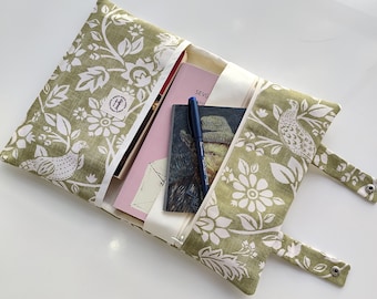 May include: A green and white floral fabric book clutch with a zipper closure and two fabric straps. The clutch is open and contains a pink book, a notebook, and a pen.