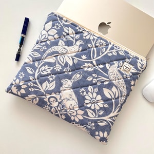 Op de afbeelding: Blauwe gewatteerde laptoptas met een bloemen- en dierenprint. Het ontwerp toont witte konijnen, vogels en bloemen tegen een blauwe achtergrond. De hoes heeft een ritssluiting en is ontworpen voor een laptop.