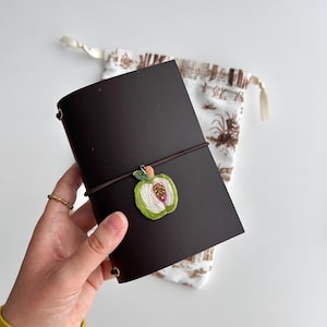 Könnte beinhalten: Ein dunkelbraunes Lederjournal mit einem gestickten Apfelmotiv. Das Journal wird mit einem braunen Gummiband verschlossen und hat einen kleinen, dekorativen Apfel auf der Vorderseite. Ein gemusterter Kordelzugbeutel ist im Hintergrund.