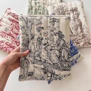 May include: Quilted fabric cases with various toile de Jouy patterns in red, green, blue, and black. The cases feature a rectangular shape with a zip closure and a stitched design. The cases are designed to protect tablets or laptops.