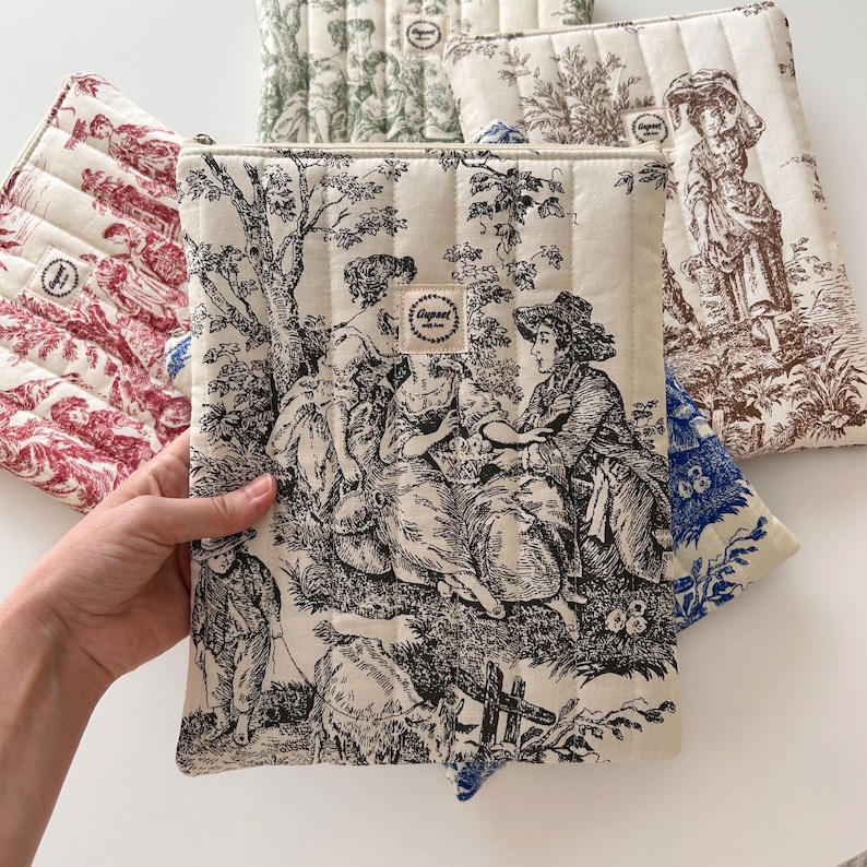 May include: Quilted fabric cases with various toile de Jouy patterns in red, green, blue, and black. The cases feature a rectangular shape with a zip closure and a stitched design. The cases are designed to protect tablets or laptops.