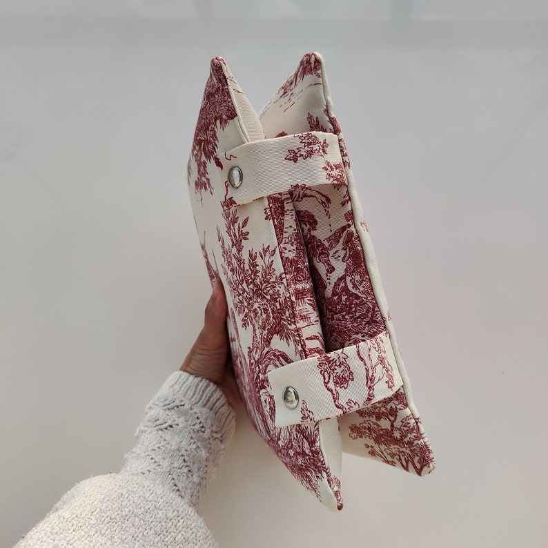 May include: A white and red toile patterned fabric clutch bag with two straps and silver metal snaps.