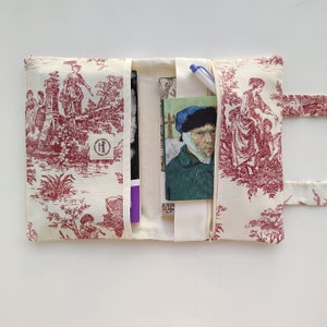 May include: A red and white toile fabric clutch with a zipper closure. The clutch has a pocket on the inside and a strap for carrying. The clutch is open to reveal a book and a pen.