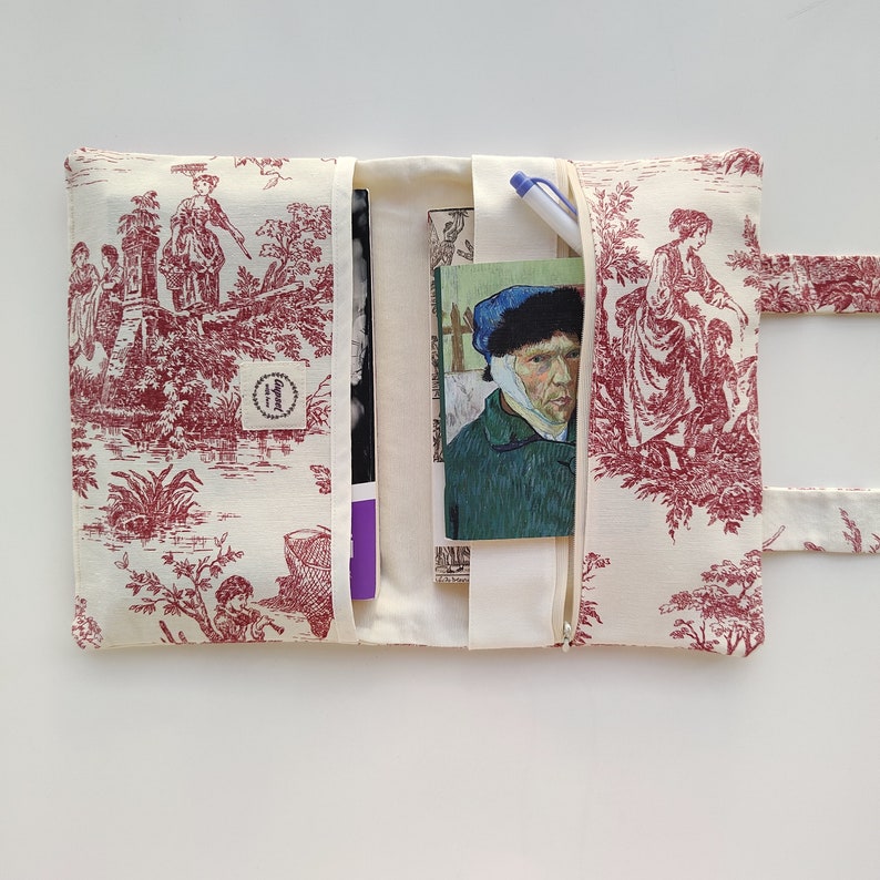 May include: A red and white toile fabric clutch with a zipper closure. The clutch has a pocket on the inside and a strap for carrying. The clutch is open to reveal a book and a pen.