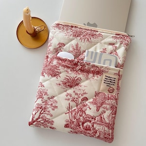 May include: Cream and red quilted laptop sleeve with a toile pattern. The sleeve has a zip closure and a front pocket containing a white mouse, a small notebook, and a tag with text. A laptop is partially visible inside.