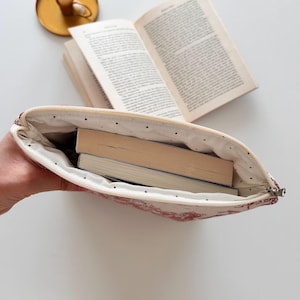May include: A cream-coloured book sleeve with a red toile pattern, holding two books. The interior is white with black polka dots. A brass candle holder and an open book are in the background.