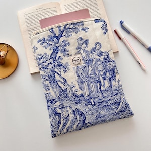 May include: A cream-coloured, quilted laptop sleeve with a blue toile design depicting figures in a rural scene. The sleeve has a zip closure and a small fabric tag with text. A book and pens are in the background.