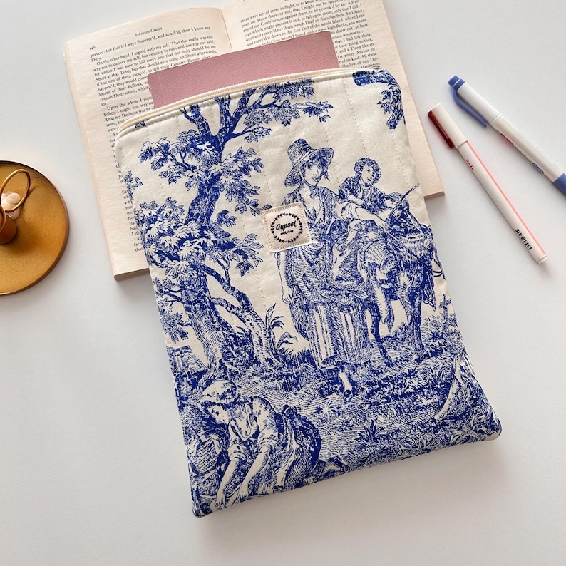May include: A cream-coloured, quilted laptop sleeve with a blue toile design depicting figures in a rural scene. The sleeve has a zip closure and a small fabric tag with text. A book and pens are in the background.