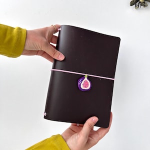 Peut inclure: Un carnet en cuir marron foncé maintenu par un élastique rose clair. Une petite breloque peinte à la main en forme de figue coupée en deux, avec une peau violette et un intérieur rose à graines, est fixée au centre de l'élastique rose.