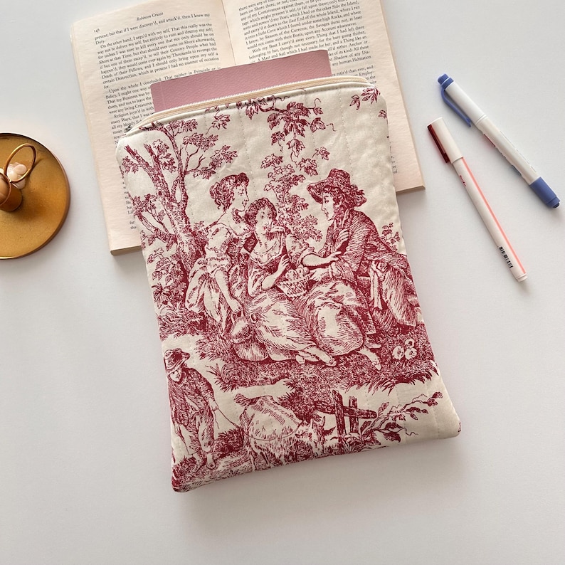 May include: A cream-coloured book sleeve with a burgundy toile pattern showing figures in a rural setting. The rectangular, padded sleeve has a zip closure. Pens and a book are visible in the background.