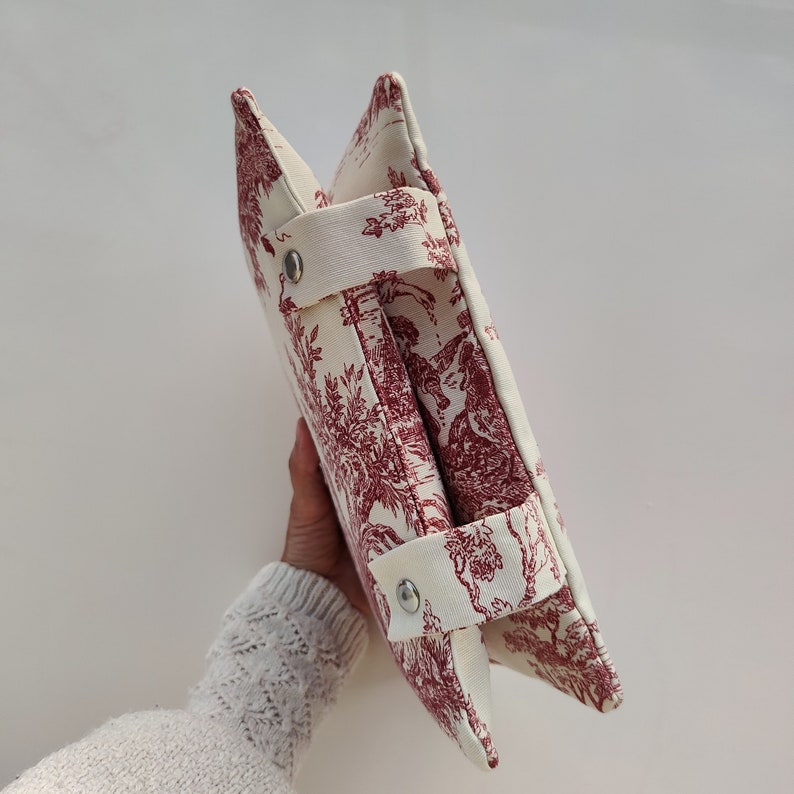 May include: A white and red toile patterned fabric storage bin with two handles and silver metal snaps. The bin is folded in half and held by a hand.