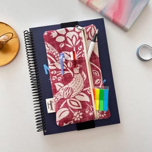 May include: A red and white patterned pencil case with a zipper, open to reveal pens and sticky notes, rests on a spiral-bound notebook. The case features a bird and floral design. The notebook is dark blue.