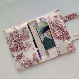May include: A white fabric clutch with a red and white toile pattern. The clutch has a zipper closure and a flap with a snap closure. The clutch is open to reveal a pen, a notebook, and a book.