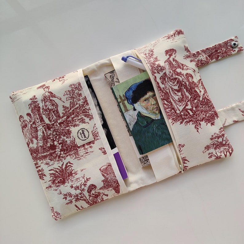 May include: A white fabric clutch with a red and white toile pattern. The clutch has a zipper closure and a flap with a snap closure. The clutch is open to reveal a pen, a notebook, and a book.