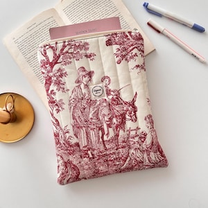May include: Cream quilted tablet sleeve with a red toile print showing figures and a donkey. The sleeve has a zip closure and a small label. A book and pens are visible in the background.