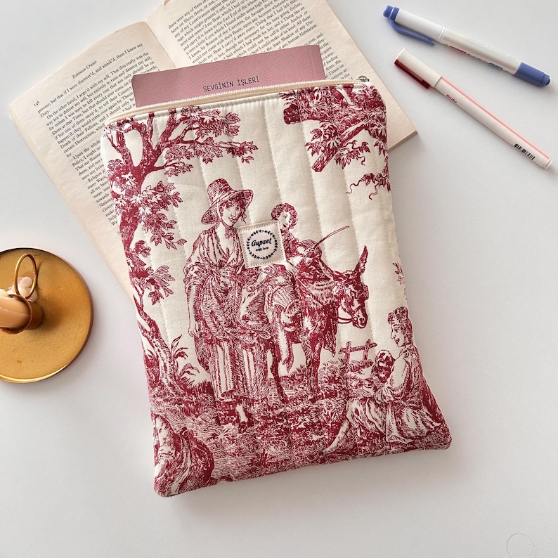 May include: Cream quilted tablet sleeve with a red toile print showing figures and a donkey. The sleeve has a zip closure and a small label. A book and pens are visible in the background.