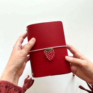 May include: A red leather journal held in place by a light pink elastic band. Attached to the elastic band is a small, embroidered strawberry charm featuring a red body with white seed details and a green leaf top, centered on the front cover.
