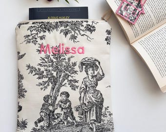 Può includere: Una custodia per libri in tela bianca con un motivo toile bianco e nero che raffigura una donna e due bambini. La custodia ha il nome "Melissa" ricamato in lettere rosa.
