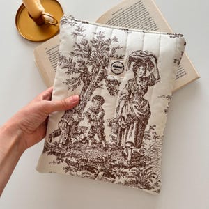 May include: Cream quilted book sleeve with a brown toile design depicting a rural scene with figures and trees. The sleeve has a zip closure and a small label that reads "Gagreet".
