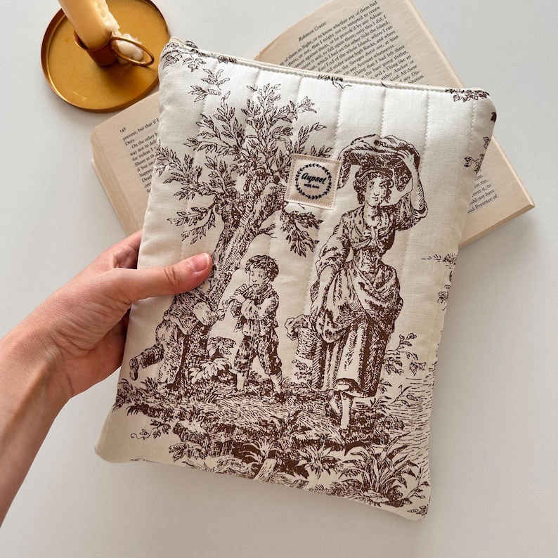 May include: Cream quilted book sleeve with a brown toile design depicting a rural scene with figures and trees. The sleeve has a zip closure and a small label that reads "Gagreet".