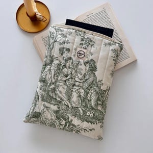 May include: Cream quilted tablet sleeve with a green pastoral toile print. The sleeve has a zip closure and a small label. A book and a candle holder are in the background.