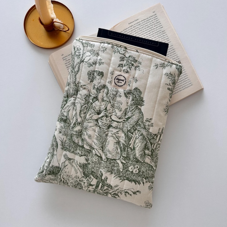 May include: Cream quilted tablet sleeve with a green pastoral toile print. The sleeve has a zip closure and a small label. A book and a candle holder are in the background.