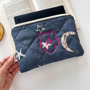 Könnte beinhalten: Eine marineblaue, gesteppte Tasche mit weißem Reißverschluss, bestickt mit Sternen, einem Mond und einem rosa Buchstaben "A". Die Tasche hat ein Rautenmuster und ein kleines Etikett mit der Aufschrift "Ciupset".