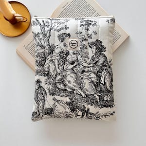 May include: A cream-coloured book sleeve with a black and white toile design depicting figures in a pastoral scene. The sleeve has a quilted texture and a small tag with the word "Gugert" printed on it. The sleeve is resting on an open book.