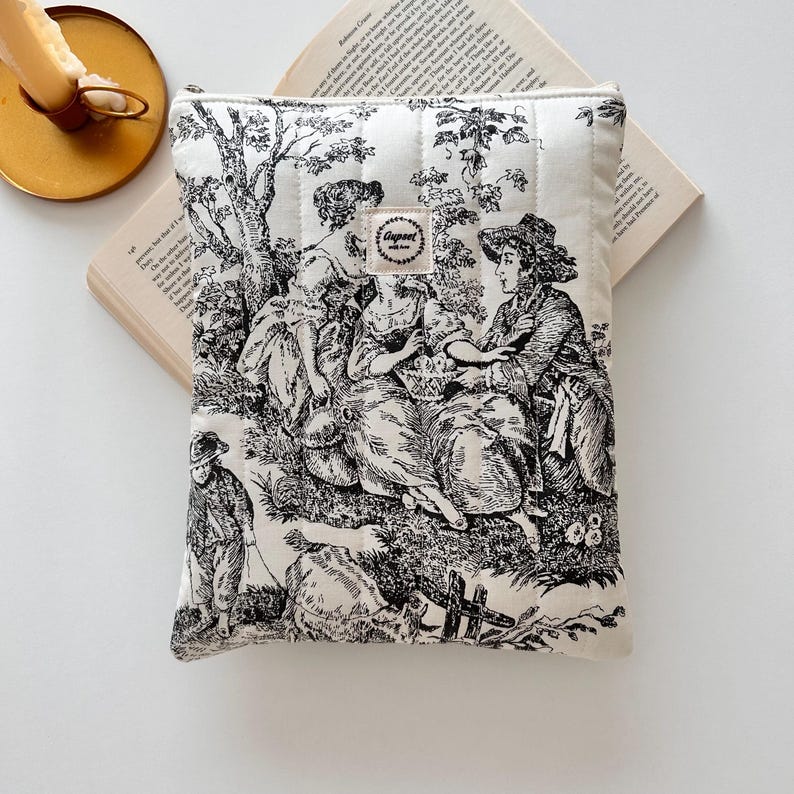 May include: A cream-coloured book sleeve with a black and white toile design depicting figures in a pastoral scene. The sleeve has a quilted texture and a small tag with the word "Gugert" printed on it. The sleeve is resting on an open book.