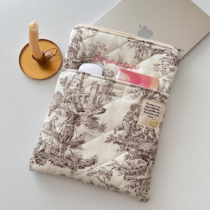 Könnte beinhalten: Cremefarbene, gesteppte Laptoptasche mit braunem Toile-de-Jouy-Muster. Die Tasche hat eine Vordertasche mit einer weißen Maus und einem rosa Gegenstand. Ein goldener Kerzenhalter mit einer brennenden Kerze befindet sich links. Reißverschluss.