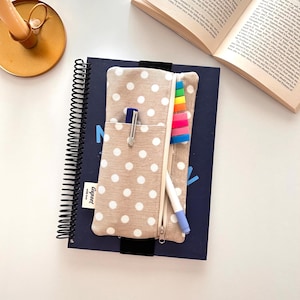 Könnte beinhalten: Ein beiges Federmäppchen mit weißen Punkten und Reißverschluss, gefüllt mit einem Stift, einem Textmarker und einem blauen Stift, liegt auf einem Spiralnotizbuch. Das Notizbuch ist marineblau, das Wort "N" ist sichtbar. Ein kleines Etikett zeigt "Gupset".