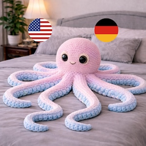 Puede incluir: Un peluche de pulpo hecho a mano en crochet, en rosa y azul. El pulpo tiene una cara sonriente con ojos grandes y redondos. Los tentáculos hacen una transición de rosa a azul. El peluche está sobre una colcha gris.