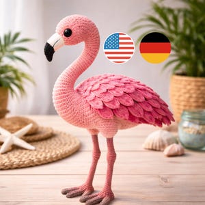 May include: A handmade pink crochet flamingo with a black and white beak, standing on two thin legs. The flamingo has a textured body and a ruffled wing design. The background includes a starfish and a woven mat.