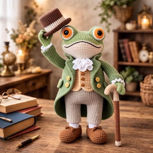Könnte beinhalten: Gehäkelte Froschfigur mit grünem Mantel, beiger Weste und grauer Hose, die einen braunen Zylinder und einen Stock hält. Der Frosch hat große orangefarbene Augen und braune Schuhe. Der Hintergrund zeigt Bücher und einen Holztisch.