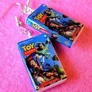 Può includere: Orecchini pendenti color argento con repliche in miniatura di VHS di Toy Story. Le cassette blu mostrano il titolo del film e i personaggi, con la scritta "IT'S LIGHT YEARS AHEAD". Su sfondo rosa.