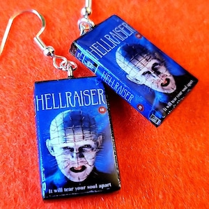 May include: A pair of silver-tone dangle earrings. Each earring features a rectangular charm with a blue background and the movie title "HELLRAISER". The image on the charm is a close-up of a character's face. The text "It will tear your soul apart" is also visible.