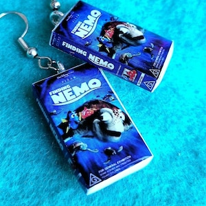 Può includere: Orecchini pendenti color argento con miniature della copertina del film "Alla ricerca di Nemo". Gli orecchini presentano il titolo e i personaggi del film su uno sfondo blu.