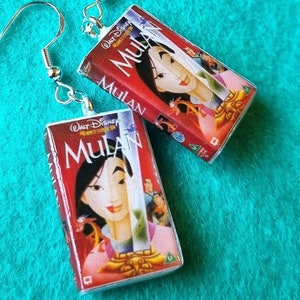 Può includere: Orecchini pendenti con repliche in miniatura delle copertine VHS del film Disney "Mulan". Gli orecchini hanno ganci color argento e le copertine sono rosse con il titolo del film e illustrazioni dei personaggi.