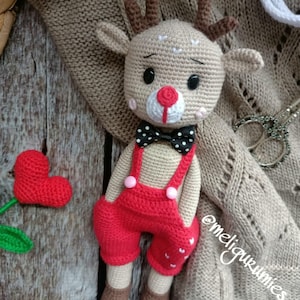 Puede incluir: Un juguete de reno de crochet que lleva un mono rojo con una pajarita de lunares negros y blancos. El reno tiene la nariz roja y los ojos negros. El juguete está sentado sobre una superficie de madera marrón.