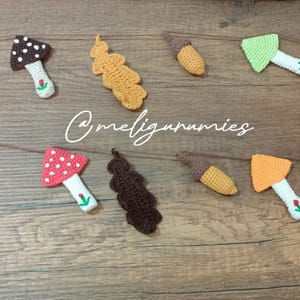 Pode incluir: Uma guirlanda de crochê feita à mão com várias formas temáticas de outono. A guirlanda inclui bolotas, cogumelos e folhas em tons de amarelo, verde, castanho e vermelho. A guirlanda é enfiada em um cordão castanho e é exibida em uma superfície de madeira.