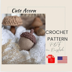 May include: A crocheted acorn with a brown cap and tan body sits on a white surface. The image includes text that reads "Cute Acorn" and "CROCHET PATTERN PDF in English". An American flag and a PDF icon are also present.