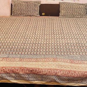 Puede incluir: Un cubrecama beige con un diseño estampado en tonos marrones, azules y rojos. El cubrecama tiene un borde con un patrón repetitivo de pequeños círculos y espirales.