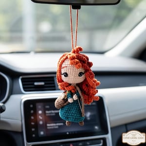 Könnte beinhalten: Handgefertigte Häkelpuppe mit roten Locken, einem türkisfarbenen Kleid und einer braunen Tasche, die an einem Faden hängt. Die Puppe hängt vor dem Innenraum eines Autos, in der Nähe des Rückspiegels. Dekorationsartikel.