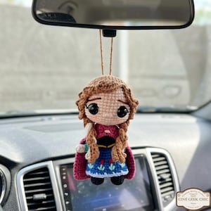 May include: A handmade crochet doll of a character with brown hair in braids, wearing a pink cape and blue dress. The doll is hanging from a string, likely as a car accessory. The doll has large black eyes and a sad expression.