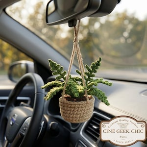 Puede incluir: Una pequeña planta colgante, tejida a ganchillo, en un coche. La planta verde está en una cesta tejida a ganchillo de color beige, suspendida por un cordón de fibra natural del espejo retrovisor. El interior del coche es visible, con el volante y el salpicadero. El texto "CINÉ GEEK CHIC" es visible.