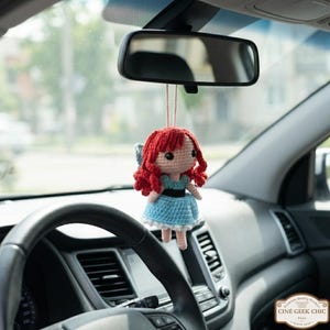 A crocheted doll with red hair and a blue dress is hanging from the rearview mirror of a car.