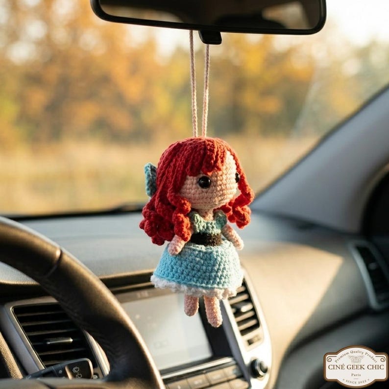 A crocheted doll with red hair and a blue dress is hanging from the rearview mirror of a car.
